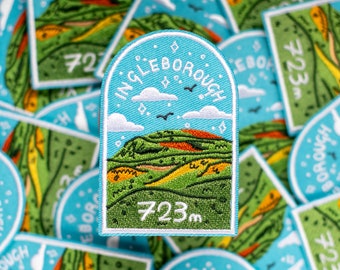 May include: A blue and green embroidered patch with the text "Ingleborough 723m" and a stylised illustration of a mountain with a red and orange peak.