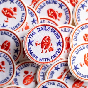 May include: A close-up of several white embroidered patches with a red outline. The patches feature a cartoon coffee bean character with a blue star and the text "The Daily Grind Easier With Coffee".