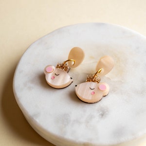 Könnte beinhalten: Ein Paar goldene und rosafarbene Ohrringe in Form einer Maus mit einer Krone oben drauf. Die Ohrringe liegen auf einer weißen Marmorfläche.