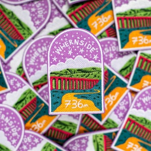 May include: A purple and green embroidered patch with the text "Whernside 736m" and a depiction of a bridge and a winding road. The patch is surrounded by other similar patches.