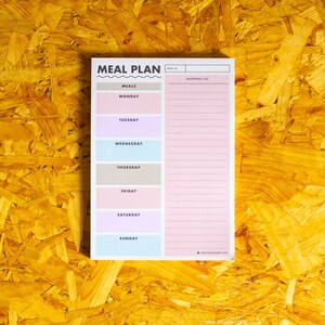 May include: A pastel-coloured meal plan notepad with the words "MEAL PLAN" at the top. The notepad has sections for each day of the week and a shopping list. The background is a textured, yellow-brown surface.