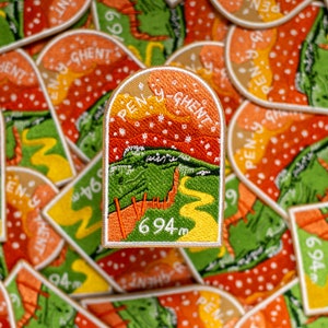 May include: A close-up of a colourful embroidered patch with the text "Pen-y-Ghent" and "694m". The patch is surrounded by other similar patches, all with different mountain names and heights.