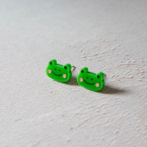 May include: A pair of green frog-shaped stud earrings with pink cheeks. The earrings are made of plastic and have a simple design.