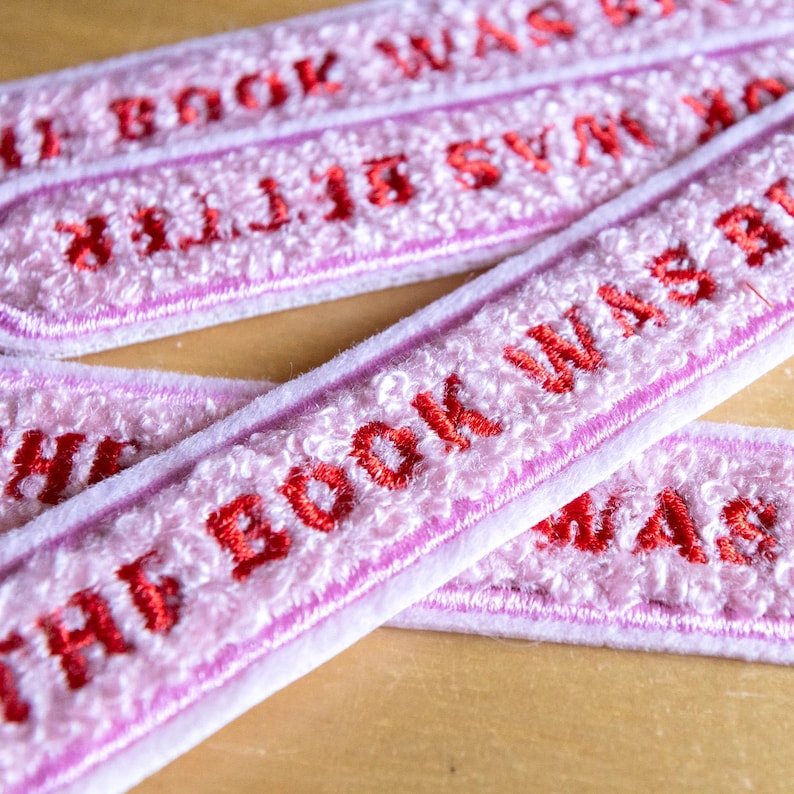 Puede incluir: Una colecci&oacute;n de parches rectangulares bordados con un fondo texturizado rosa claro y el texto THE BOOK WAS BETTER cosido en hilo rojo intenso. Cada parche tiene un borde acabado y est&aacute; dispuesto en un patr&oacute;n superpuesto.