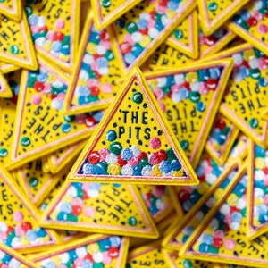 May include: A yellow triangular patch with the text "The Pits" in black lettering. The patch is filled with colorful, embroidered dots.