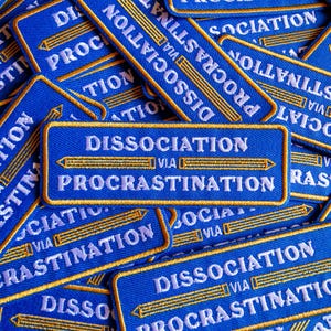 May include: Blue embroidered patches with the text "DISSOCIATION VIA PROCRASTINATION" in white and gold. The patches feature a pencil design. The patches are scattered across the image.