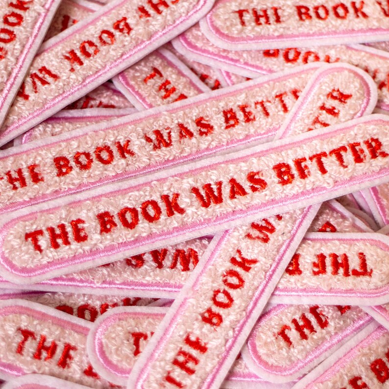Puede incluir: Una colecci&oacute;n de parches rectangulares bordados con un fondo texturizado de color rosa claro, un borde rosa y el texto THE BOOK WAS BETTER cosido en letras may&uacute;sculas rojas y negritas en el centro de cada parche individual.