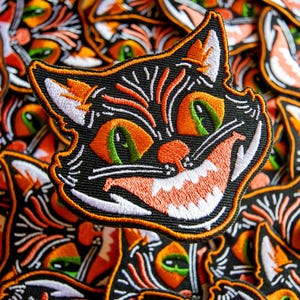 May include: Embroidered patch of a black cat's face with orange and white detailing. The cat has green eyes, orange eyebrows, and a wide, toothy grin. The patch is outlined in black and orange, ideal for Halloween or cat enthusiasts.