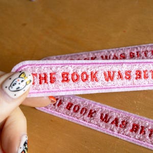 Puede incluir: Dos parches rectangulares bordados con un fondo texturizado rosa claro y un borde rosa m&aacute;s oscuro. El texto THE BOOK WAS BETTER est&aacute; bordado en letras may&uacute;sculas rojas llamativas en el centro de cada parche.