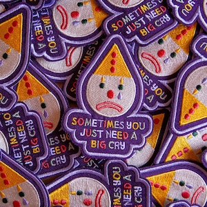 Könnte beinhalten: Gestickte Aufnäher mit einem traurigen Clown-Design. Die Aufnäher haben einen lila Rand, einen gelben Clownhut und den Text "Sometimes You Just Need A Big Cry". Der Clown hat eine rote Nase und eine Träne.
