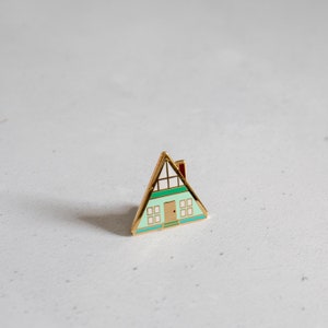 Könnte beinhalten: Eine goldfarbene Emaille-Anstecknadel in Form eines Hauses mit grünem Dach, weißen Fenstern und einem roten Schornstein.
