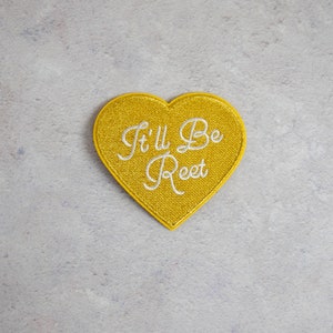 May include: A gold heart-shaped patch with the text "It'll Be Reet" embroidered in white.