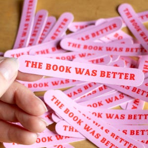 Puede incluir: Un conjunto de marcapáginas rosas, de forma oblonga, con el texto rojo "THE BOOK WAS BETTER". Los marcapáginas están dispersos, con uno sostenido en una mano. El tema sugiere un producto literario o relacionado con libros.