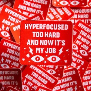 May include: A collection of red square stickers with white text that reads "HYPERFOCUSED TOO HARD AND NOW IT'S MY JOB." The stickers also feature eye and lightning bolt graphics. The stickers are scattered.