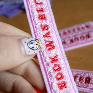 Puede incluir: Un parche rectangular bordado en color rosa claro con una superficie texturizada similar a la tela de toalla. El parche presenta el texto bordado en rojo "BOOKWORM" orientado verticalmente, enmarcado por un borde fino cosido en rosa oscuro.
