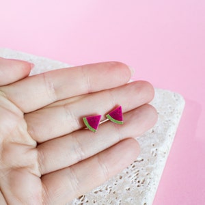 May include: A pair of watermelon slice stud earrings. The earrings are pink with green rind and black seeds.