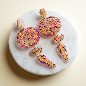 Könnte beinhalten: Ein Paar Hängeohrringe mit pinken, violetten und goldenen Glitzerakzenten. Die Ohrringe haben ein großes kreisförmiges Design mit einem goldenen Mandala-Muster und einen kleineren tropfenförmigen Anhänger.