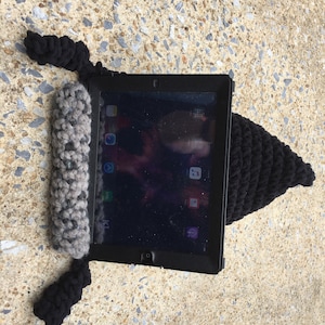 May include: A black crocheted hat with a grey and white crocheted strap. The hat is designed to hold a tablet or iPad.