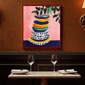 May include: A table setting with two place settings, each with a white napkin and a wine glass. The table is made of marble and has a gold base. A framed painting of a stack of colorful plates with a green plant hanging over them is on the wall behind the table.