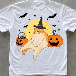May include: A white t-shirt featuring a graphic of an orange cat wearing a black witch hat, sitting between two jack-o'-lanterns. One pumpkin holds fish bones, and the background includes a yellow moon, black stars, and flying bats.