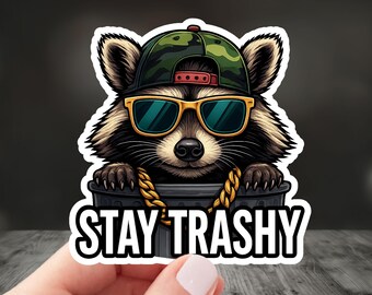 May include: A sticker featuring a raccoon wearing a camouflage baseball cap, gold-rimmed sunglasses, and a gold chain. The raccoon is peeking out of a trash can, with the words "STAY TRASHY" in white block letters.