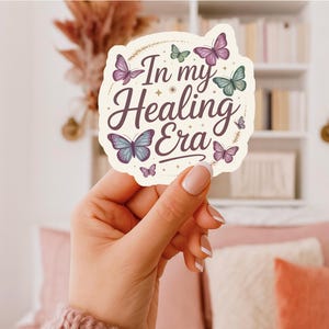 May include: A sticker with the text "In my Healing Era" in a decorative font, surrounded by butterflies in shades of purple, blue, and green. The sticker has a cream-colored background and a whimsical design.