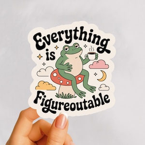May include: A die-cut sticker featuring a cartoon frog sitting on a mushroom and holding a coffee cup. The sticker has the words "Everything is Figureoutable" in a retro font, with stars, clouds, and a crescent moon design.
