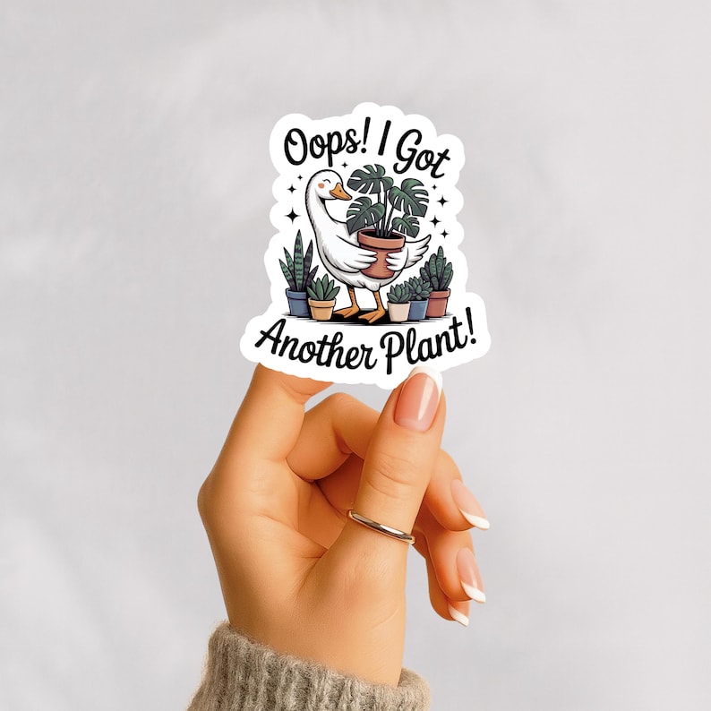 A hand holding a funny plant sticker that says " Opps! I got another plant!" It has a goose carring a plant on its back and is surrounded by many other plants. Sticker has a white background and the sticker and the hand is placed on a light gray background.