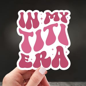 May include: Pink sticker with the text "In My Titi Era" in a retro font with white outlines and glitter accents.
