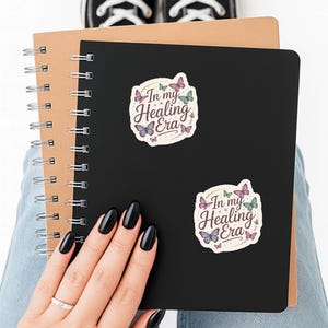 May include: A black spiral notebook with two stickers that read "In my Healing Era" with butterfly illustrations. The notebook is held by a person wearing blue jeans and black sneakers.