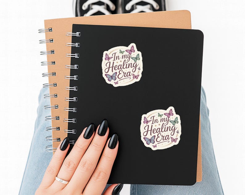 May include: A black spiral notebook with two stickers that read "In my Healing Era" with butterfly illustrations. The notebook is held by a person wearing blue jeans and black sneakers.