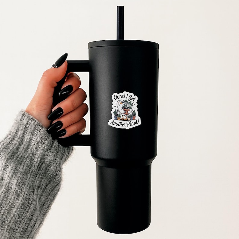 May include: Black insulated tumbler with a handle and a black straw. A white sticker on the tumbler reads "Oops! I Got Another Plant!" with a cartoon goose and potted plants. The tumbler is designed to keep beverages cold or hot.