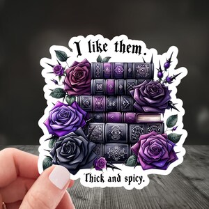 An image of a hand holding a romance reader-themed sticker that says " I like them thick and spicy".  The sticker has a stack of books in purple and dark purple colors, and purple and dark gray roses are arranged in between the books with thorns.