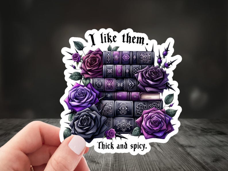 An image of a hand holding a romance reader-themed sticker that says " I like them thick and spicy".  The sticker has a stack of books in purple and dark purple colors, and purple and dark gray roses are arranged in between the books with thorns.