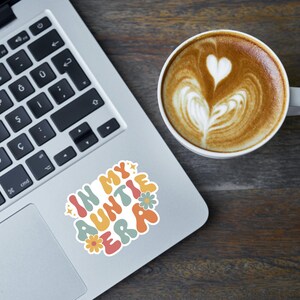 May include: A laptop computer with a sticker that says "In My Auntie Era" in colorful letters with flowers. The sticker is on the silver laptop next to a white coffee mug with latte art.