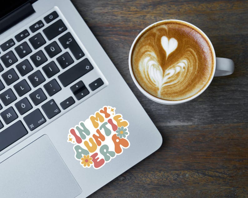 May include: A laptop computer with a sticker that says "In My Auntie Era" in colorful letters with flowers. The sticker is on the silver laptop next to a white coffee mug with latte art.