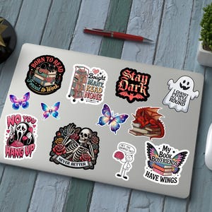 May include: A laptop covered in colorful stickers with various designs, including a ghost, butterflies, a skeleton, and text that reads "Born to Read" and "My Book Boyfriends Have Wings." A red pen and a computer mouse are also visible.
