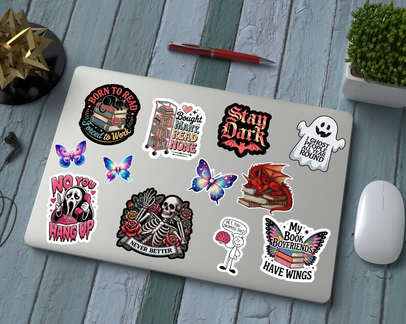 May include: A laptop covered in colorful stickers with various designs, including a ghost, butterflies, a skeleton, and text that reads "Born to Read" and "My Book Boyfriends Have Wings." A red pen and a computer mouse are also visible.