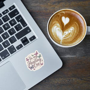 May include: A laptop with a sticker that reads "In my Healing Era" with butterfly illustrations. A cup of coffee with latte art in the shape of a heart sits next to the laptop on a wooden surface.