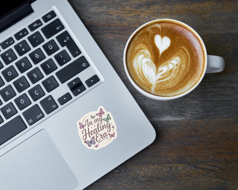 May include: A laptop with a sticker that reads "In my Healing Era" with butterfly illustrations. A cup of coffee with latte art in the shape of a heart sits next to the laptop on a wooden surface.