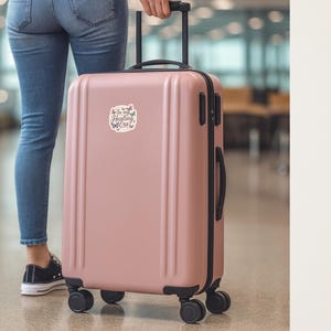 May include: A dusty rose rolling suitcase with black accents and wheels. A sticker on the front reads "In my Healing Era" with butterfly graphics. The suitcase is being pulled through an airport.
