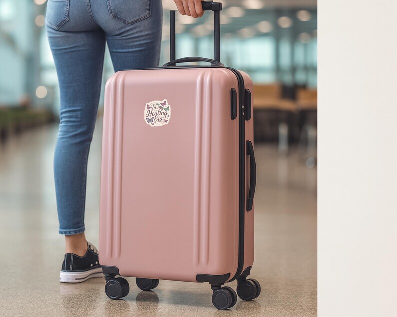 May include: A dusty rose rolling suitcase with black accents and wheels. A sticker on the front reads "In my Healing Era" with butterfly graphics. The suitcase is being pulled through an airport.
