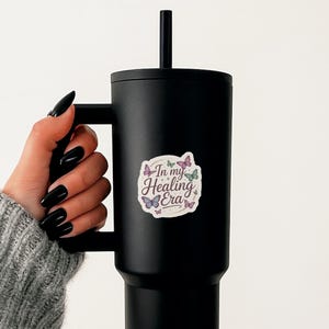May include: A black tumbler with a handle and a black straw. A sticker with the text "In my Healing Era" and butterfly illustrations is affixed to the tumbler. The tumbler is held by a person wearing a gray sweater.