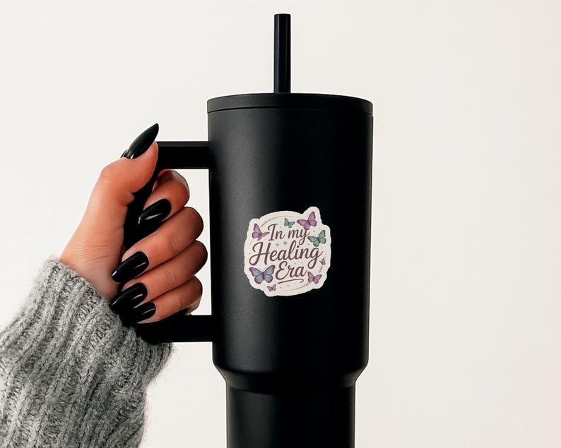 May include: A black tumbler with a handle and a black straw. A sticker with the text "In my Healing Era" and butterfly illustrations is affixed to the tumbler. The tumbler is held by a person wearing a gray sweater.