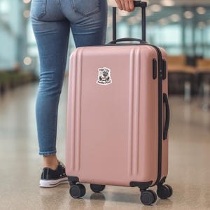 May include: A blush pink rolling suitcase with a black handle and wheels. A sticker on the suitcase reads "Oops! I Got Another Planet!" in a whimsical font, with a cartoon alien design.