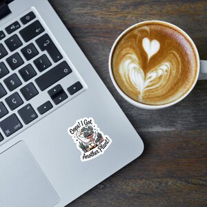 May include: A laptop with a sticker that reads "Oops! I Got Another Plant!" next to a cup of coffee with latte art. The coffee has a heart design on top. The laptop is on a dark wooden surface.