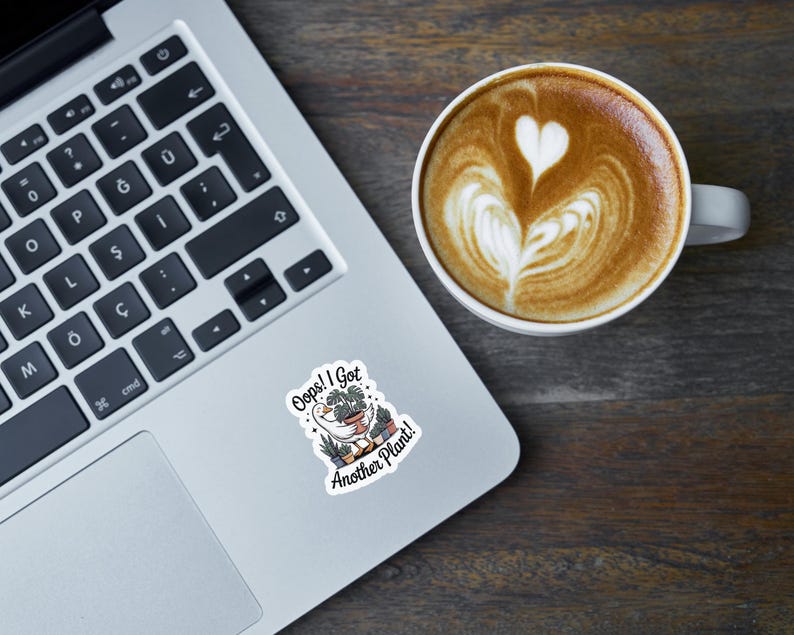 May include: A laptop with a sticker that reads "Oops! I Got Another Plant!" next to a cup of coffee with latte art. The coffee has a heart design on top. The laptop is on a dark wooden surface.