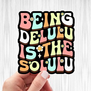 May include: A colorful sticker with the text "BEING DELULU IS THE SOLULU" in a retro font. The sticker has a black outline and is decorated with small yellow flowers.