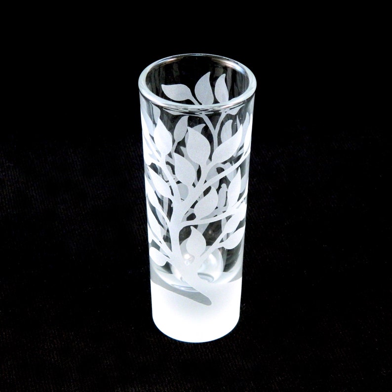 May include: A clear glass shot glass with a frosted bottom. The glass is etched with a white design of leaves and branches.