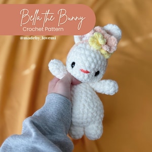 May include: A white crocheted bunny with a pink and yellow flower headband. The bunny has a red nose and black eyes. The bunny is being held by a hand wearing a gray sweatshirt. The text "Bella the Bunny Crochet Pattern @madeby_lovemi" is on a pink background.